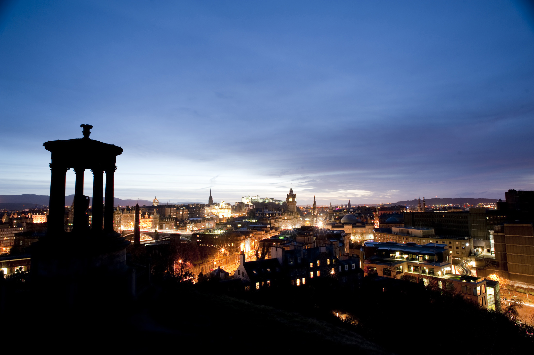 an image of Silhouette of the Dugald Stewart Monument on carlton hill with a panoramic view of edinbrugh at night behind