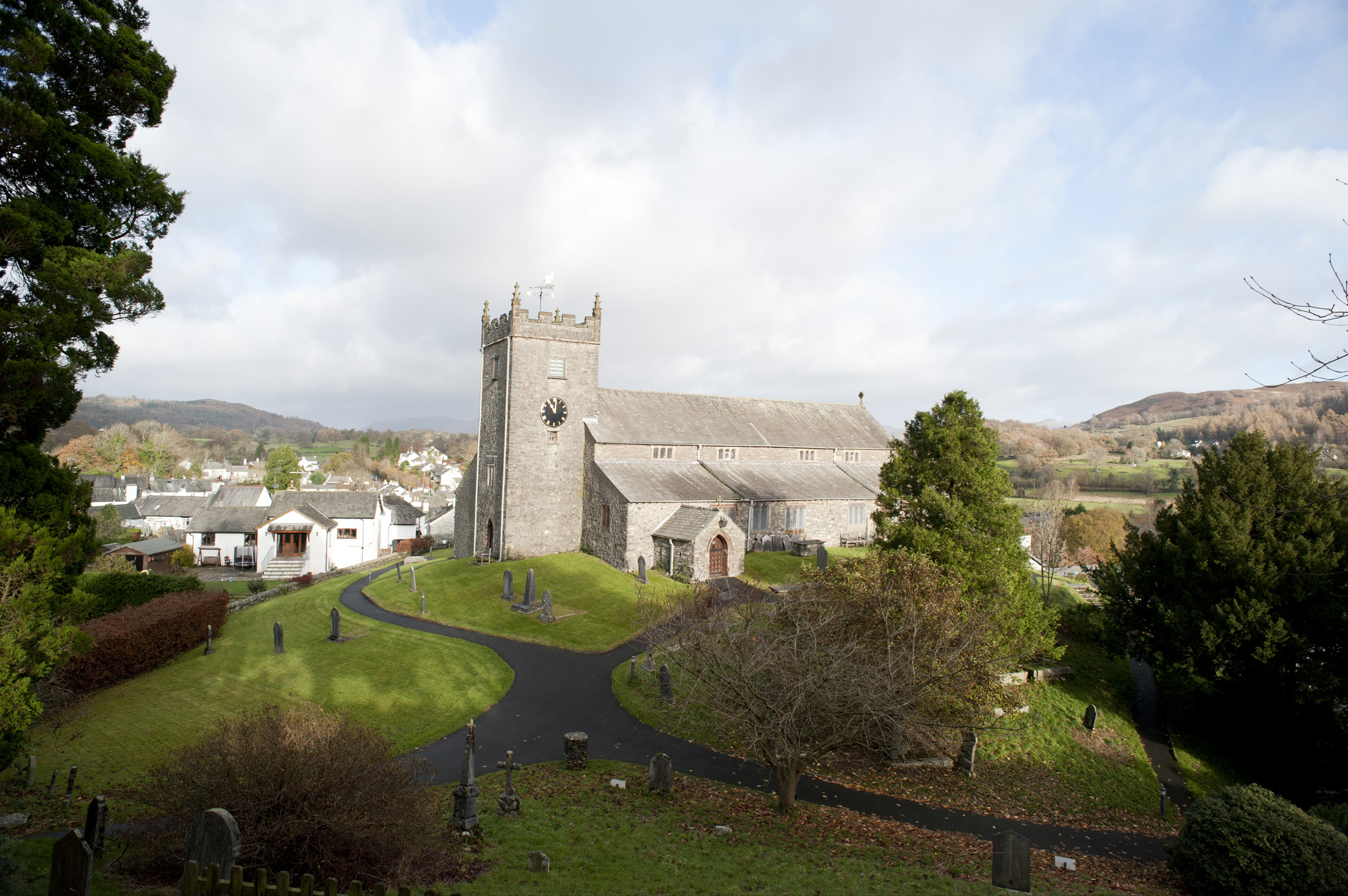 an image of Picturesque view between trees of the Hawkshead stone church and village in the Lake District in Cumbria with rolling hills behind
