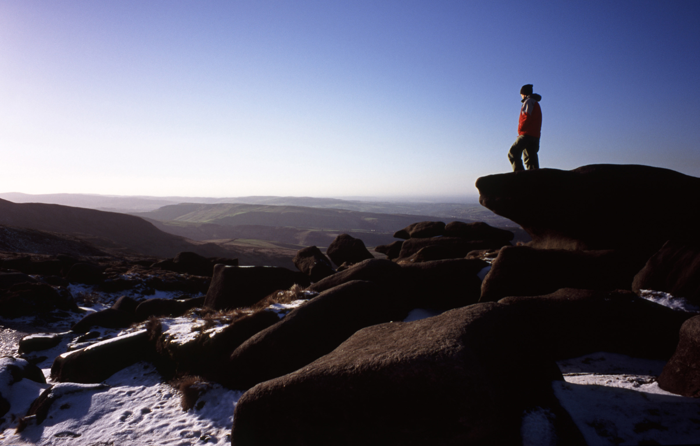 an image of Man hiking on the Kinder Scout Plateau in winter standing on a rocky outcrop admiring the view over the moorlands in the Derbyshire Peak District