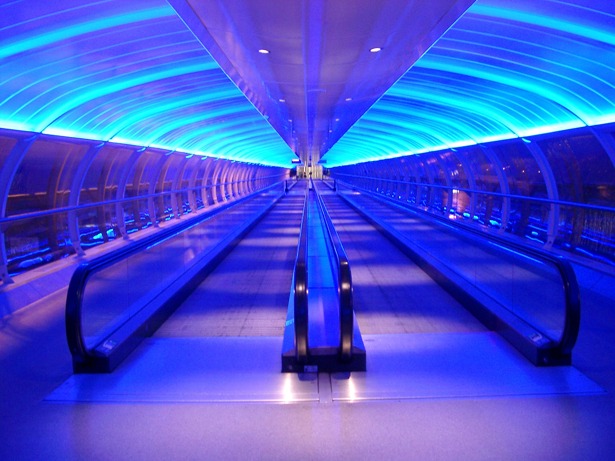an image of Interior of an airport walkway with two central moving travelators or passenger conveyor belts under a curved ceiling illuminated with blue lighting