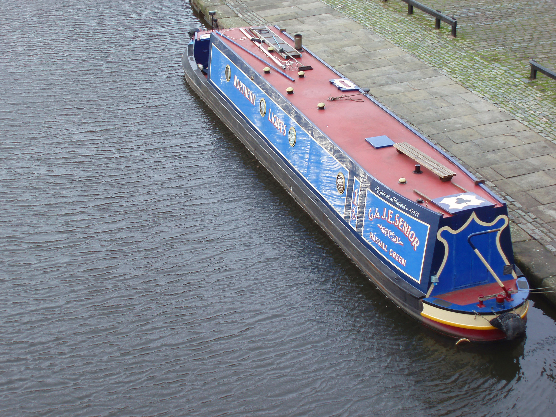 an image of High angle view of a traditional English canal narrow boat with advertising signs on the side moored at the side of the canal alongside a paved walkway