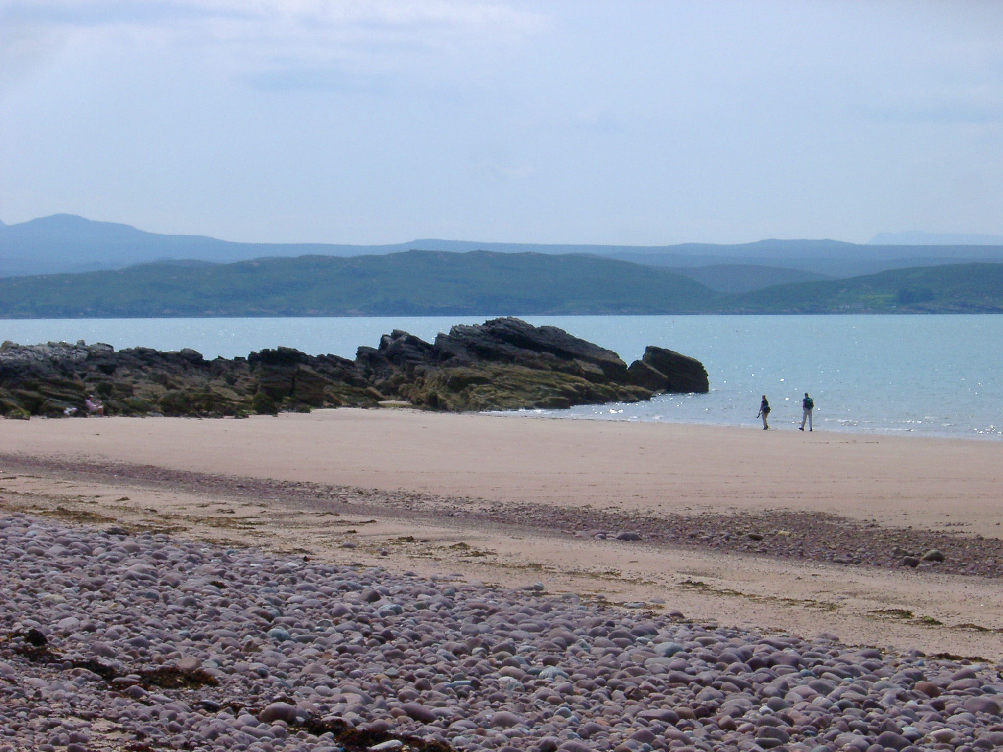 an image of Sandy beach on the shores of Lock Gairloch in the Scottish highlands with people walking along the waters edge in the sunshine