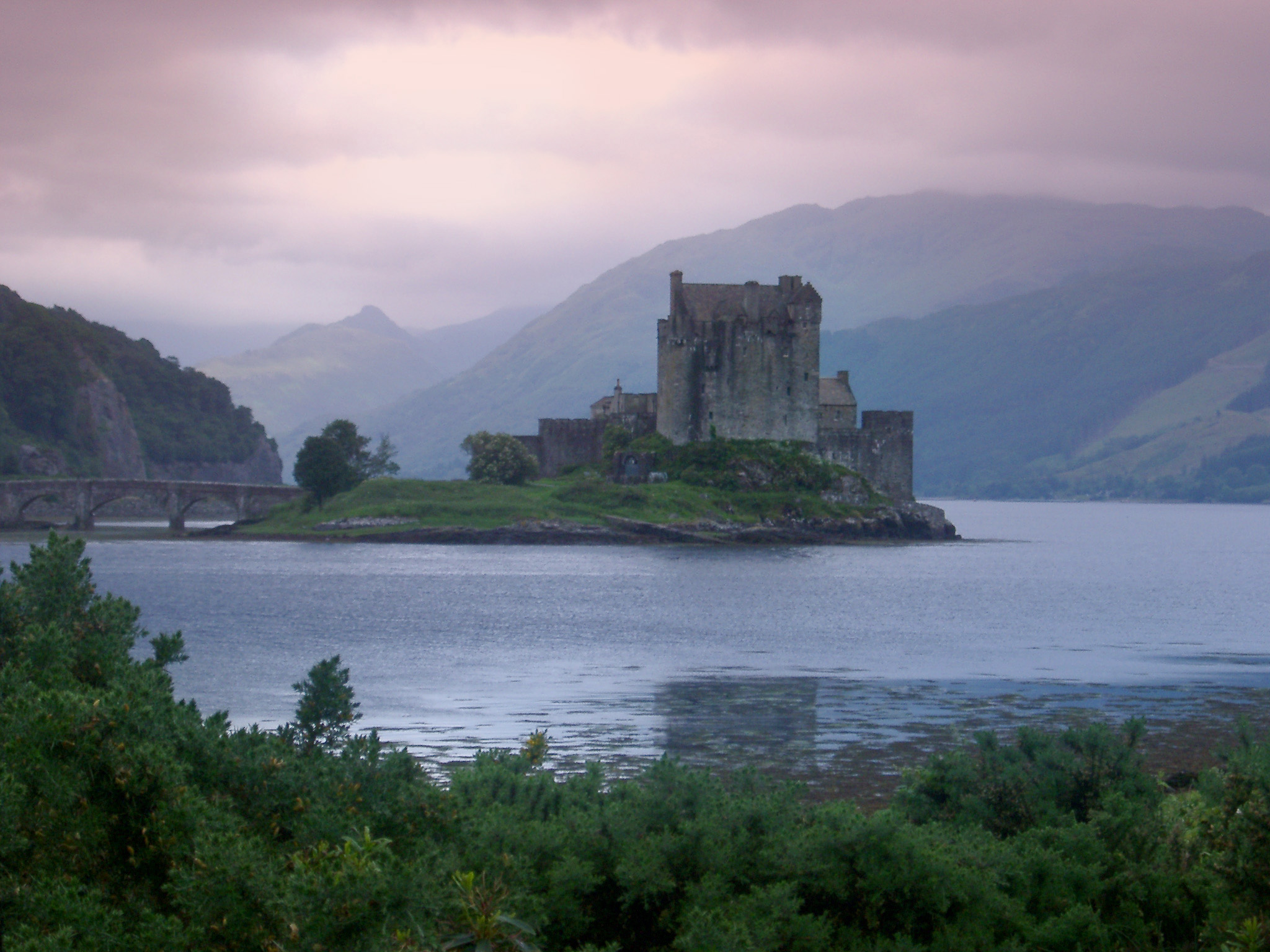 an image of Scenic atmospheric panoramic view of Eilean Donan Castle, Scotland standing on its island in the centre of Loch Duich on a calm cloudy day with a feint mist and purple tint to the sky