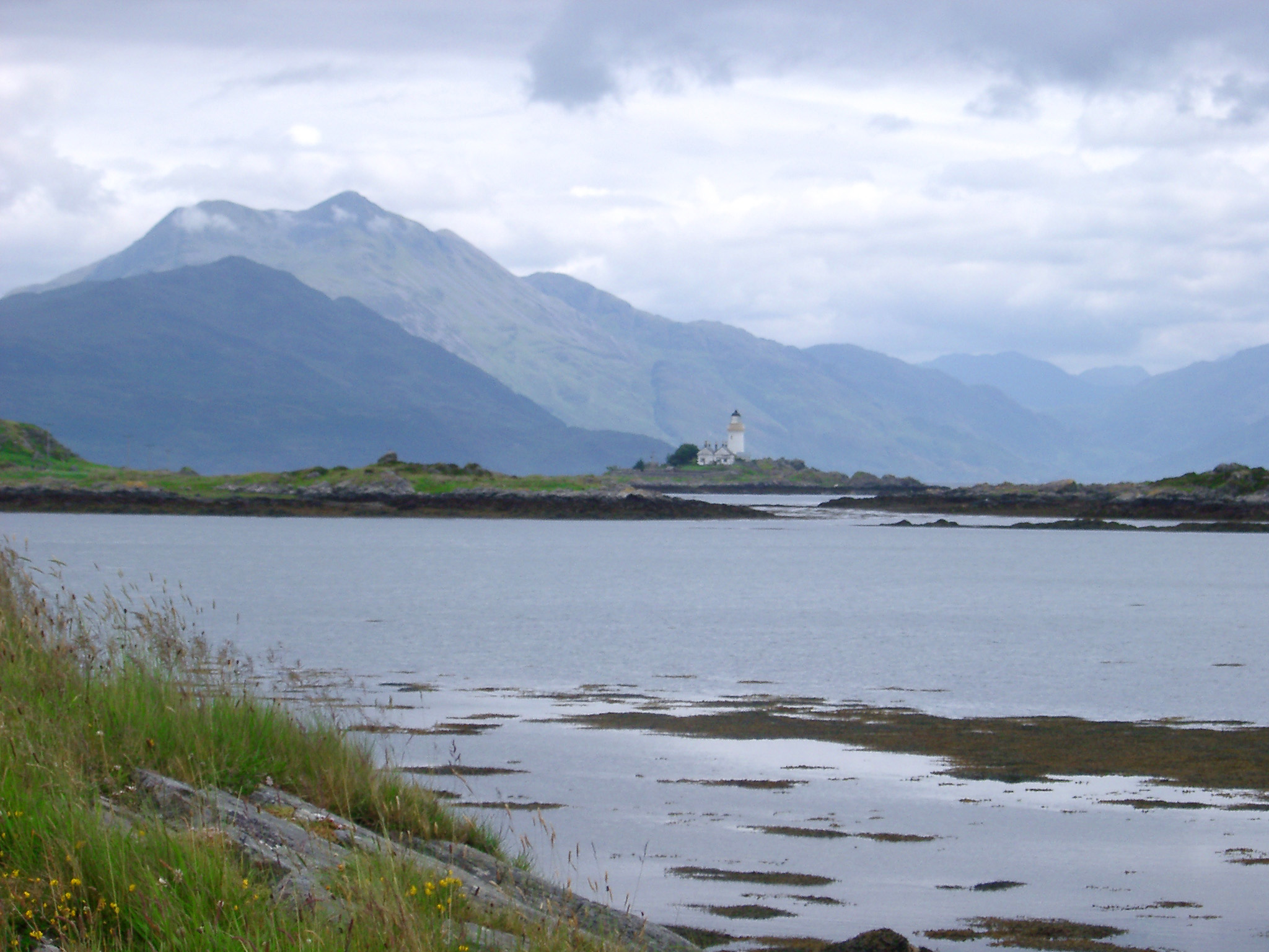 an image of Scenic view across the water of the Isle of Skye and its lighthouse below mountain peaks on a sunny cloudy day