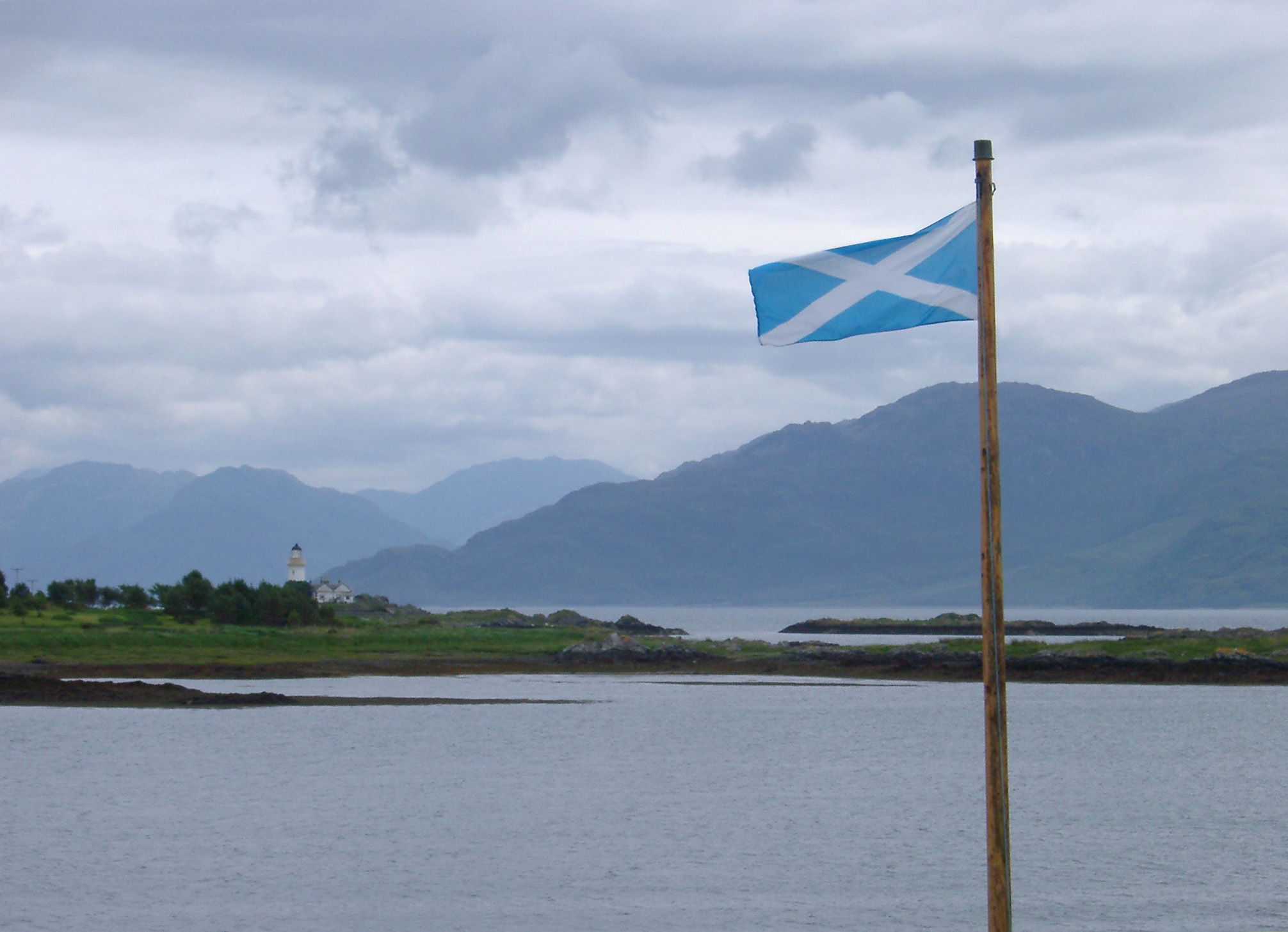 an image of National flag of Scotland flying on a flagpole overlooking a placid loch on a cold cloudy day