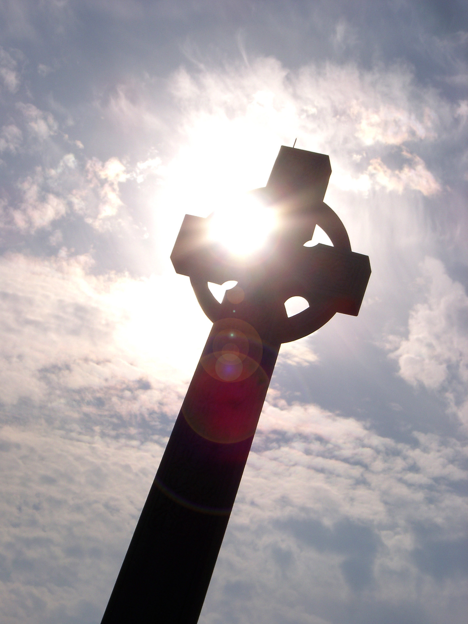 an image of Low angle view looking up at a stone Celtic cross with sun flare silhouetted against a cloudy blue sky