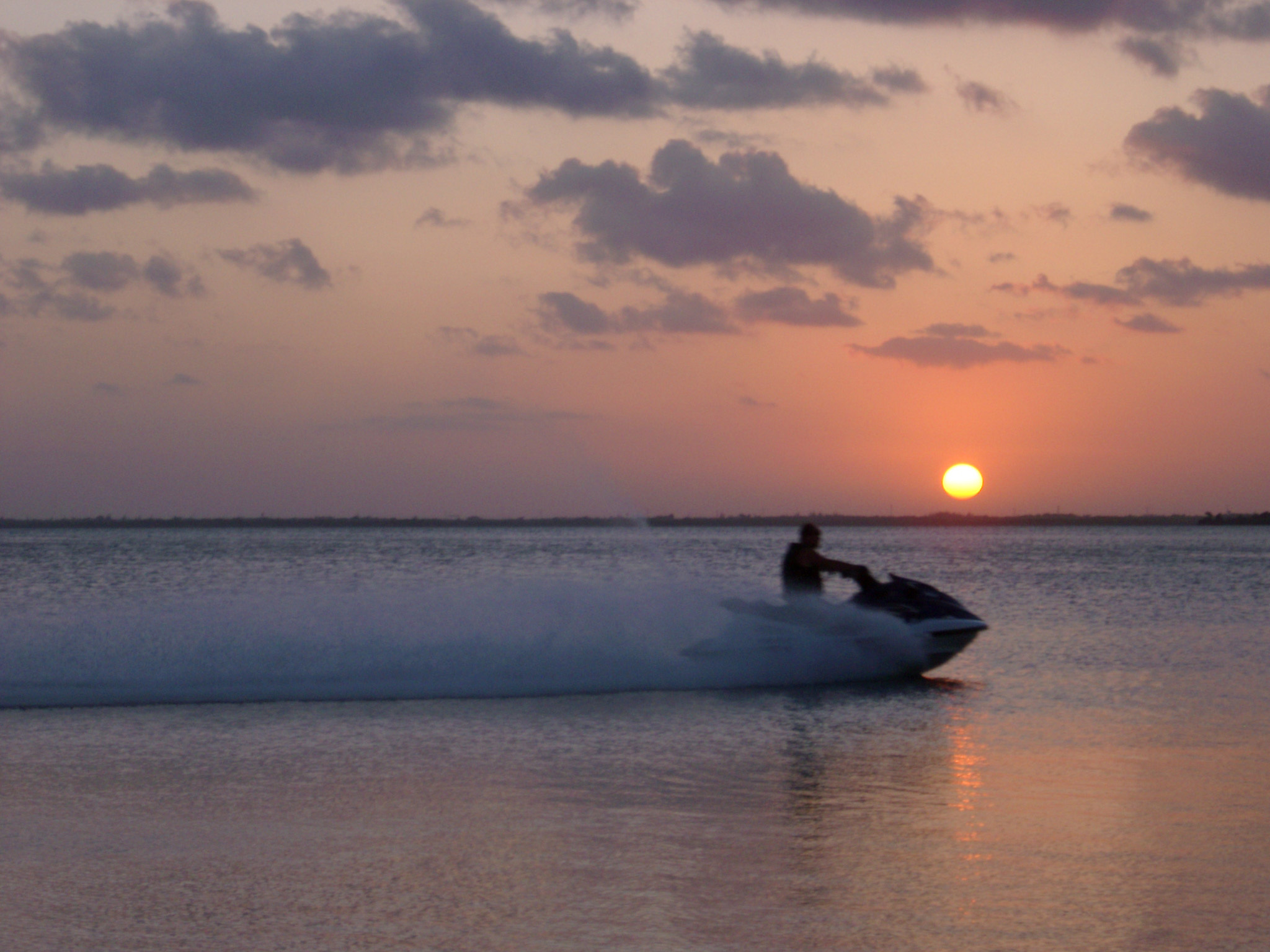 an image of Jet ski at sunset throwing up a wake and spray of turbulent water as it speeds through the ocean silhouetted against a colorful cloudy sky