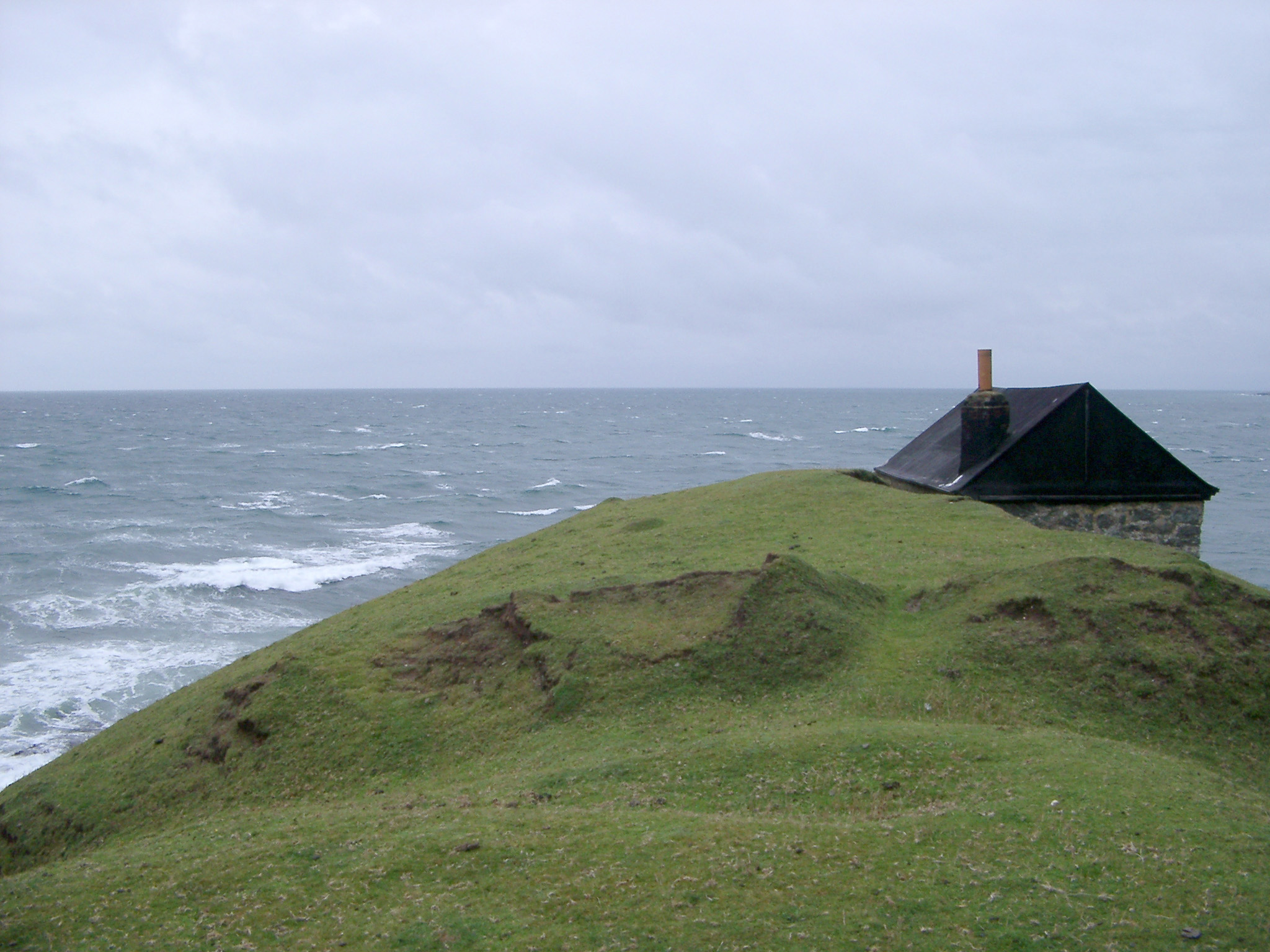 an image of Seascape with the roof of a remote cottage or shed just visible above the headland overlooking a stormy sea on an overcast day