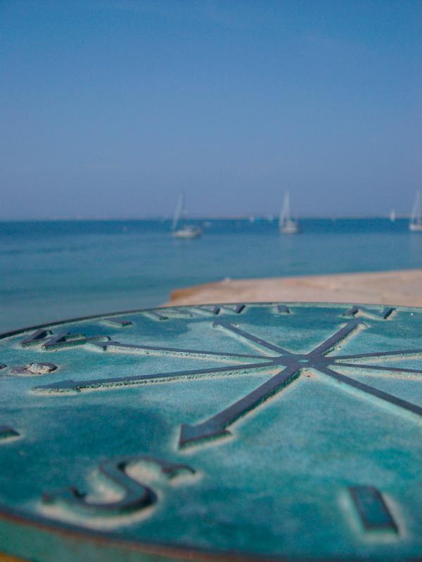 Oblique low angle view of a brass marine compass and compass points for navigation overlooking a beach with boats in the far distance