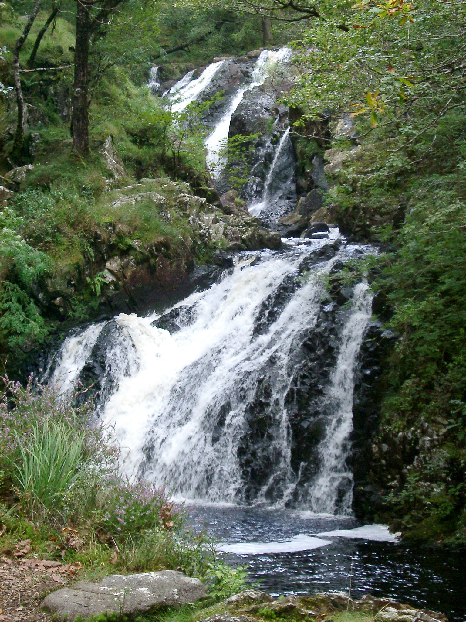 an image of Double waterfall cascading down a mountain slope over rocks into a pool below through lush green woodland in a scenic landscape