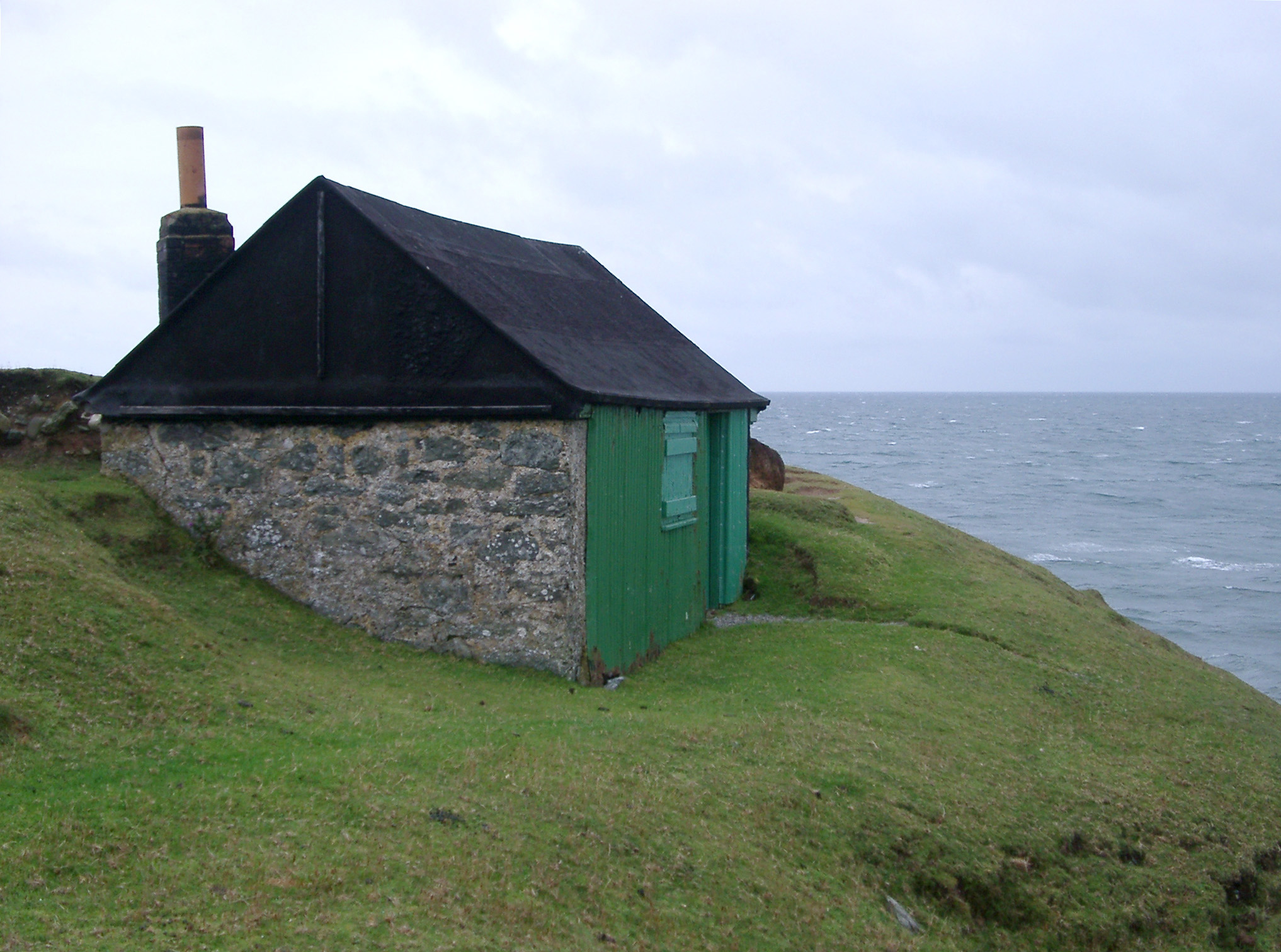 an image of Stone shed or hut with a green facade on a headland overlooking the ocean providing shelter from the coastal weather