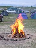 Campfire burning in a campsite amongst rows of small tents and parked cars