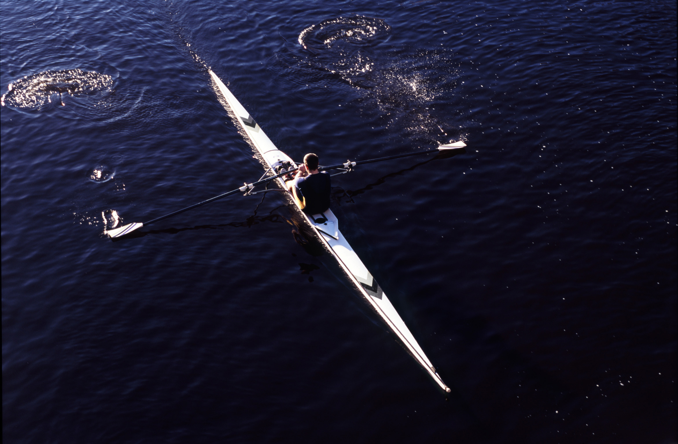 an image of Overhead view of a man practising sculling in a single boat rowing along a river with water splash off the oars