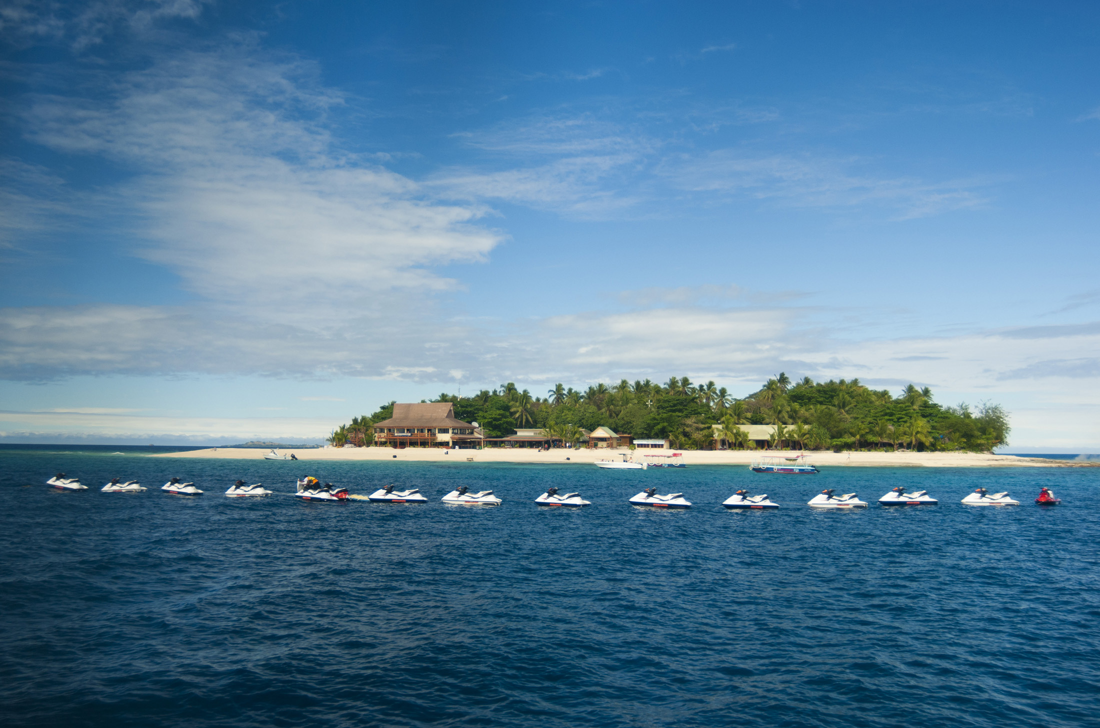 an image of View across ocean of a line of jet skis moored in front of Beachcomber Island, Fiji with its small resort
