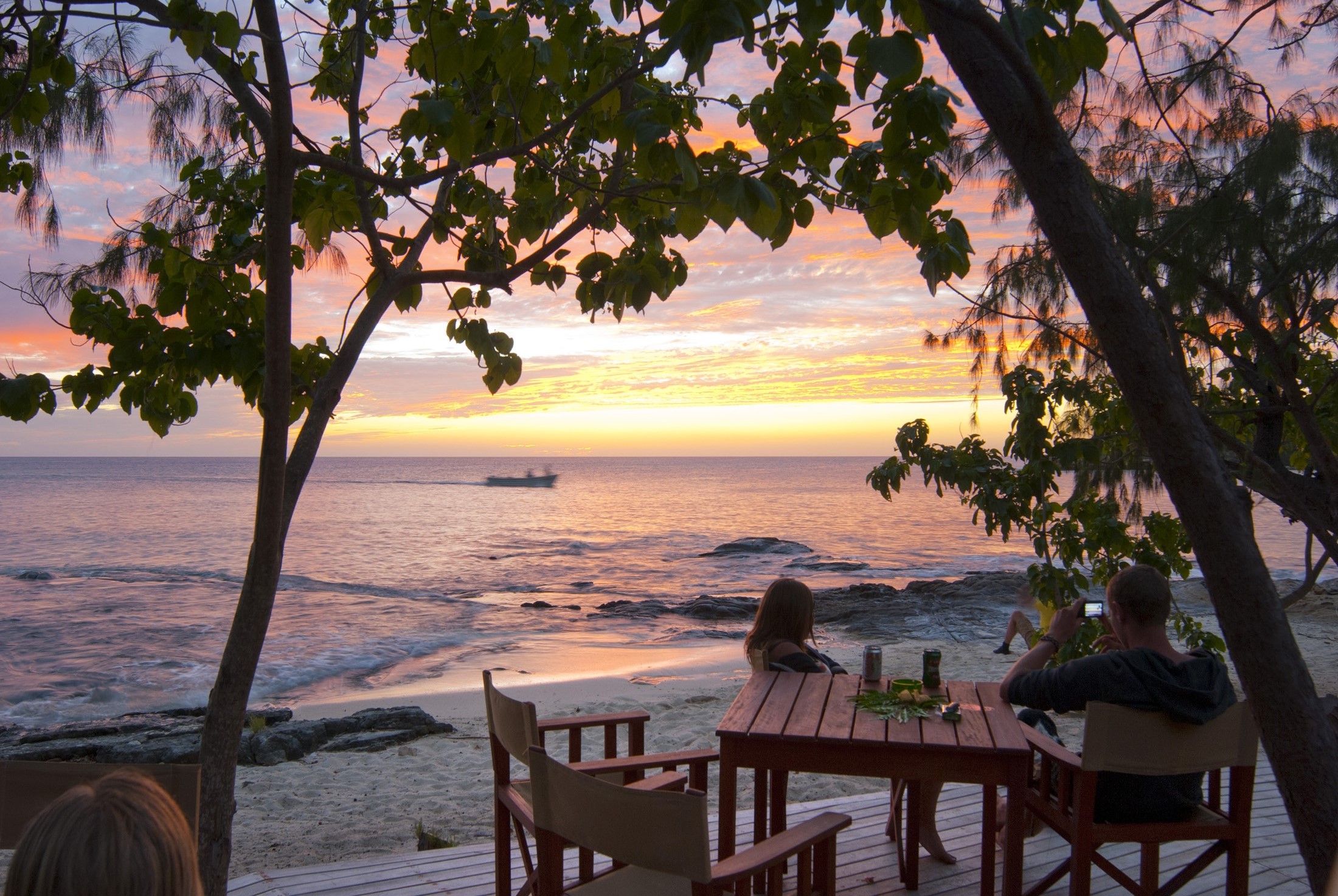 an image of People seated at a table on a deck overlooking the beach and the ocean enjoying a beautiful tropical sunset