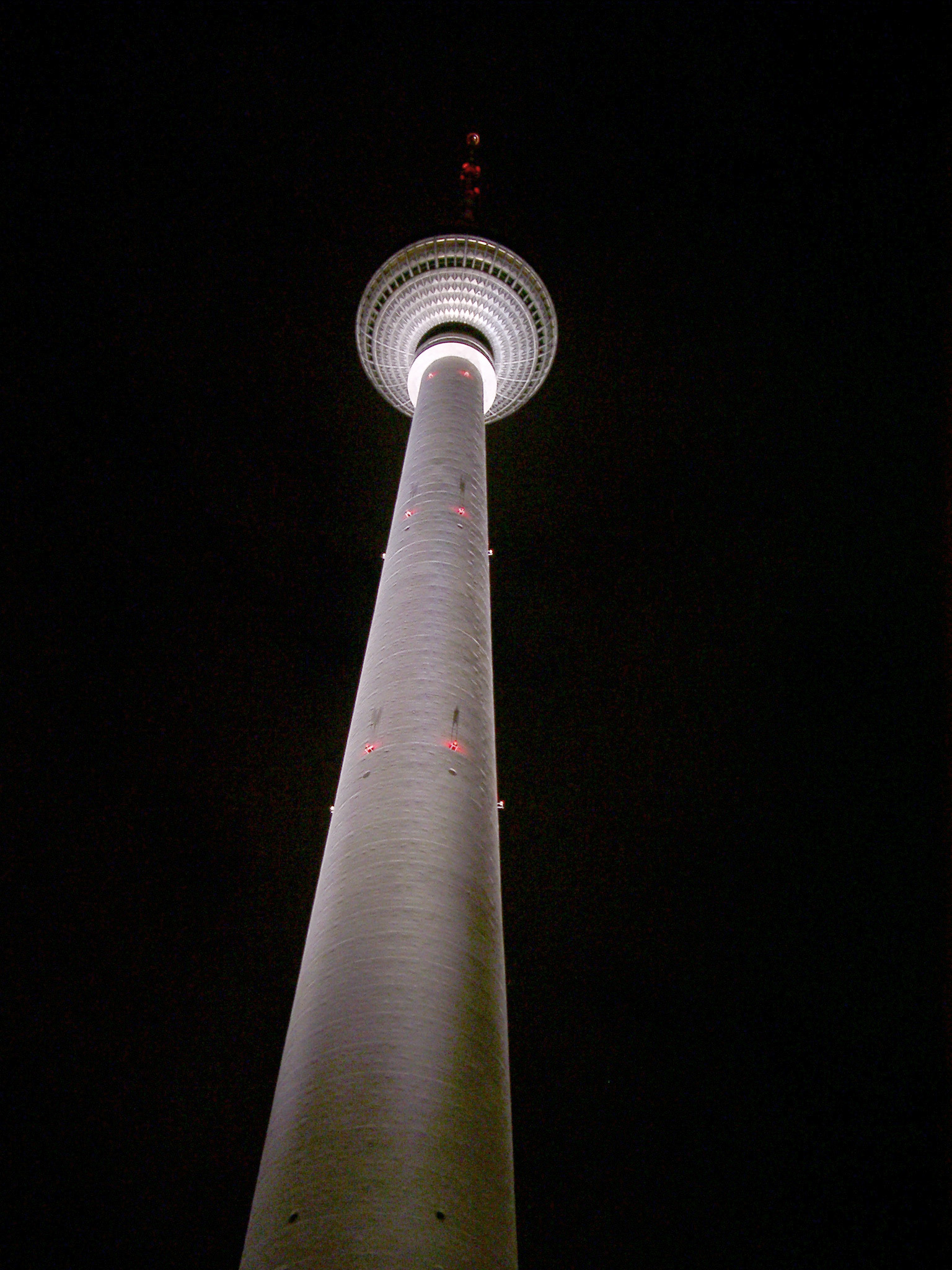an image of The Berliner Fernsehturm TV Tower at night viewed from the foot looking up to the sphere housing the restaurants and observation centre, illuminated against a dark night sky