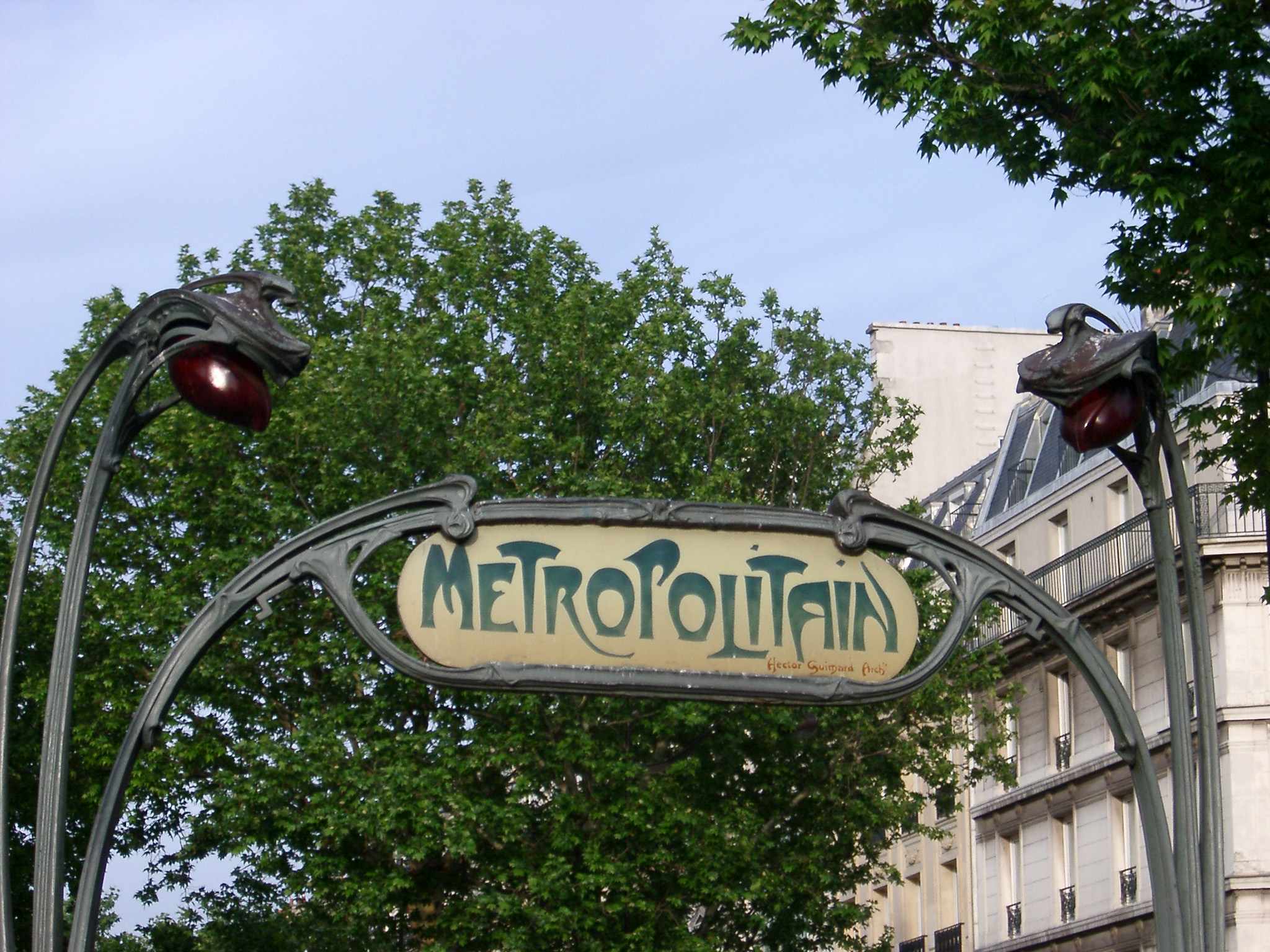 an image of Art Deco Paris Metro sign with a typical swirling design and the word - Metropolitain - against urban buildings and trees