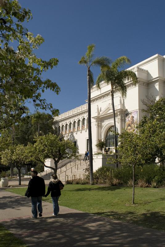 Random Tourists and Vintage Architectural Structures at Balboa Park San Diego. Surrounded by Green Trees and Plants on Blue Gray Sky Background.