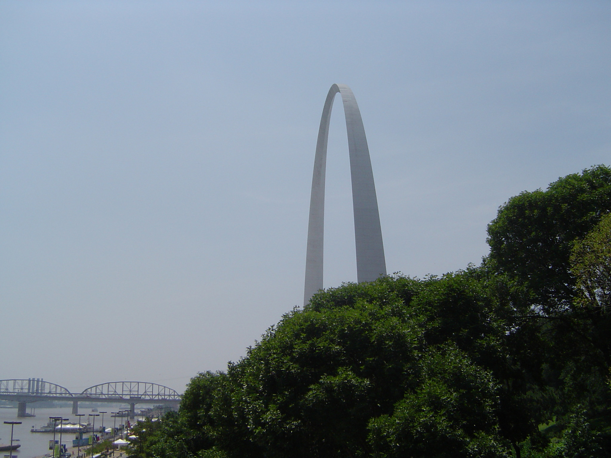an image of Famous St. Louis Gate Way Arch in Side View Surrounded by Green Trees on Light Blue Gray Sky Background.