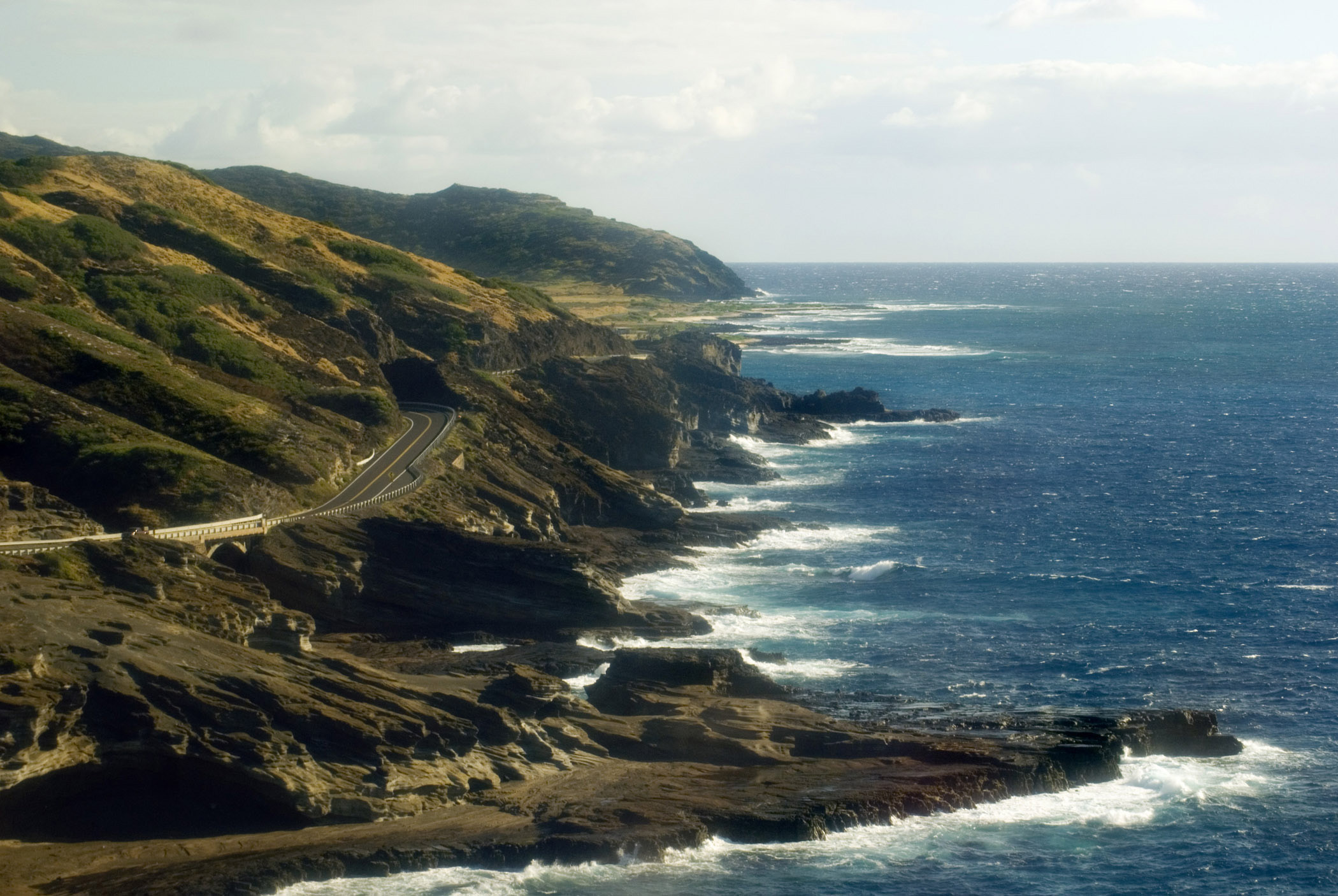 an image of Shore Line Along Kalanianaole Highway, Oahu in Aerial View. Captured with Blue Water Sea and Green Huge Mountains on Lighter Blue Sky Background.