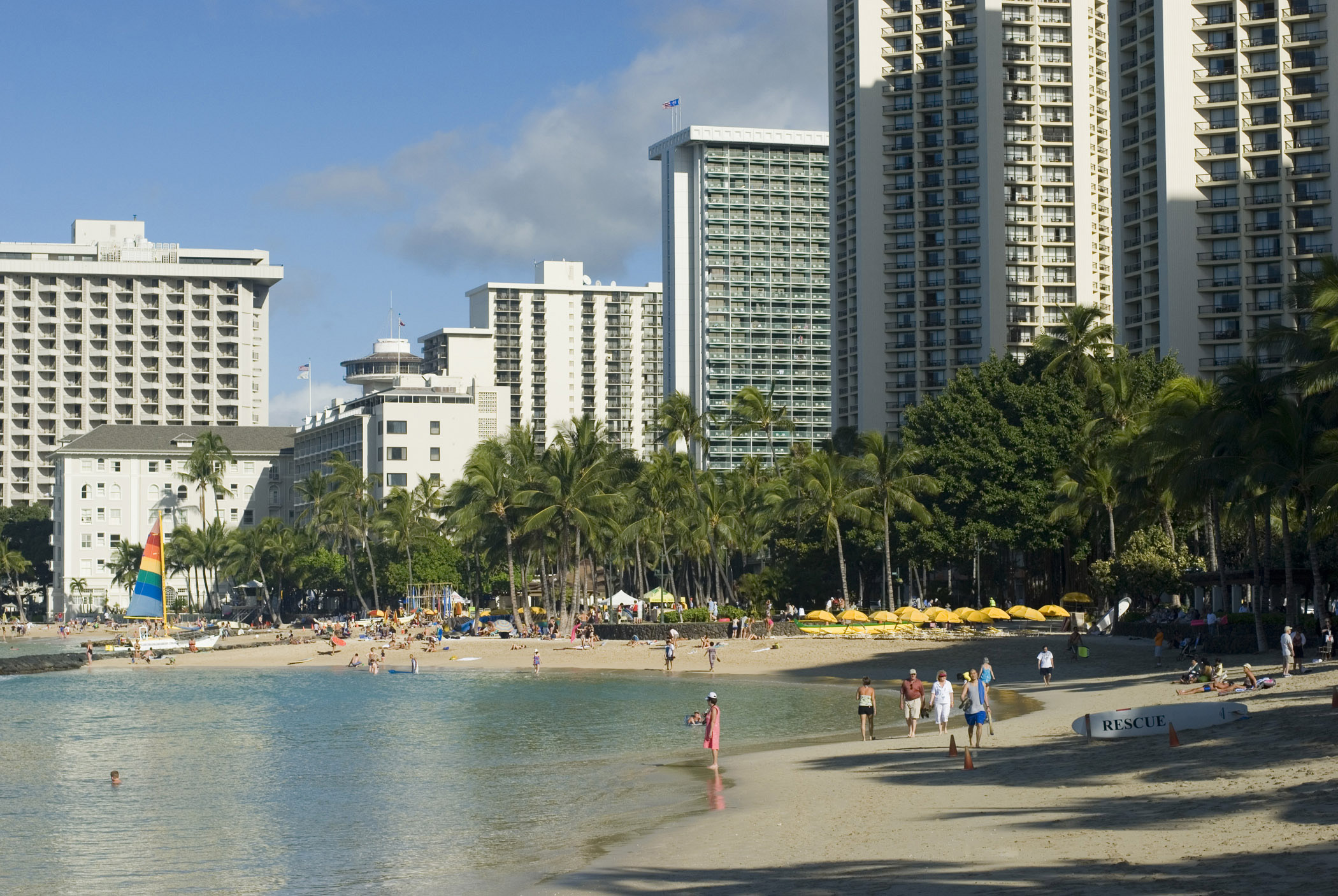 an image of Random Tourists at Beautiful Seashore of Waikiki Beach. Captured with Huge Buildings at Beachfront.