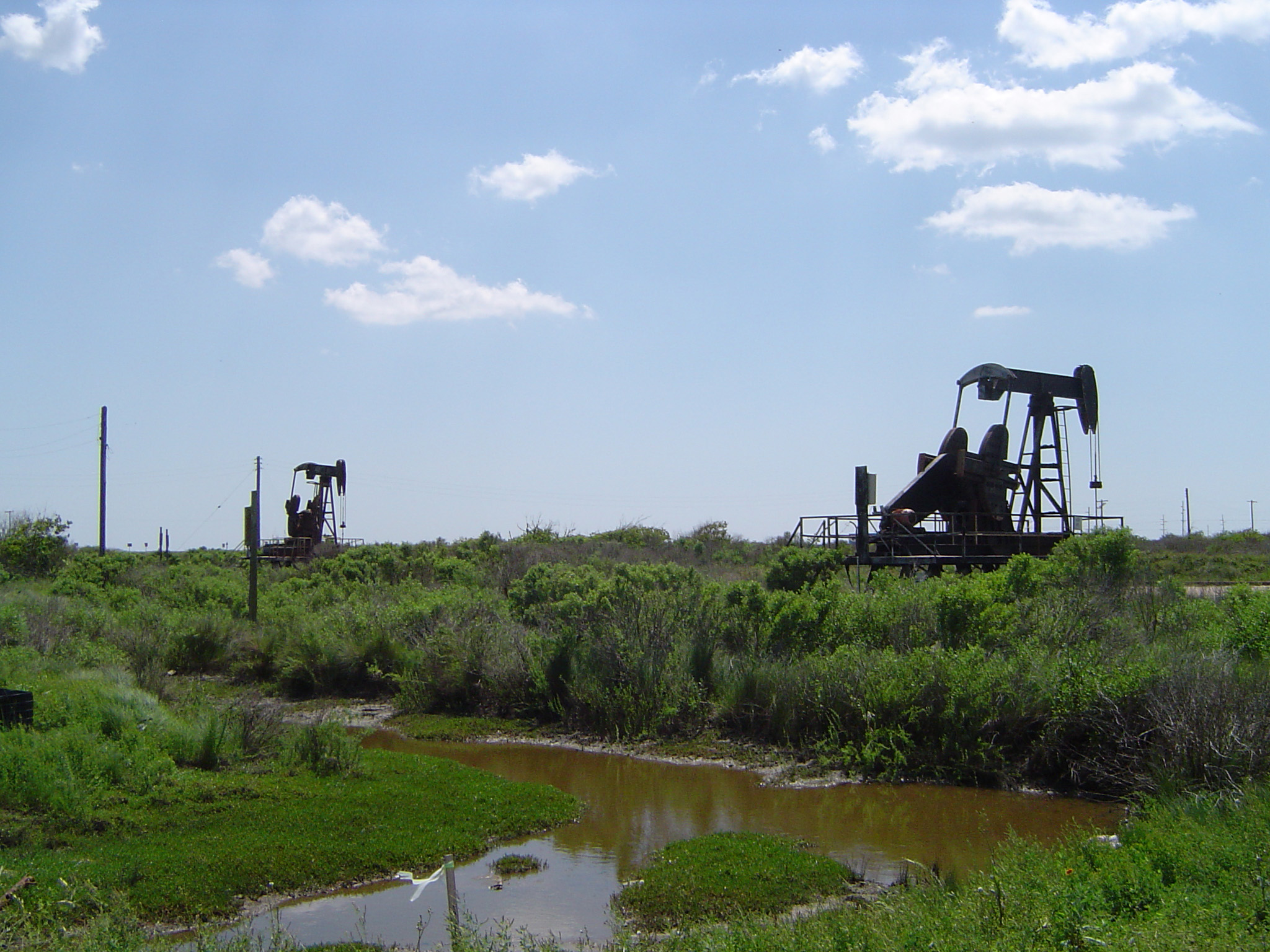 an image of Nodding Donkey, Nickname of Oil Well Drill Surrounded by Green Grasses and Flowing Water at Texas. Isolated on Light Blue Sky Background.