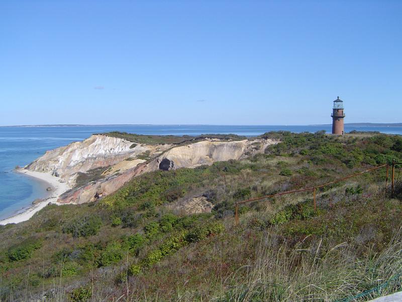 Grassy Ocean Cliff with Lighthouse Afar in Extensive View. Isolated on Light Blue Sky Background.