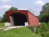 Close up Covered Bridge Surrounded by Green Grasses and Tall Trees on Lighter Blue Sky Background.