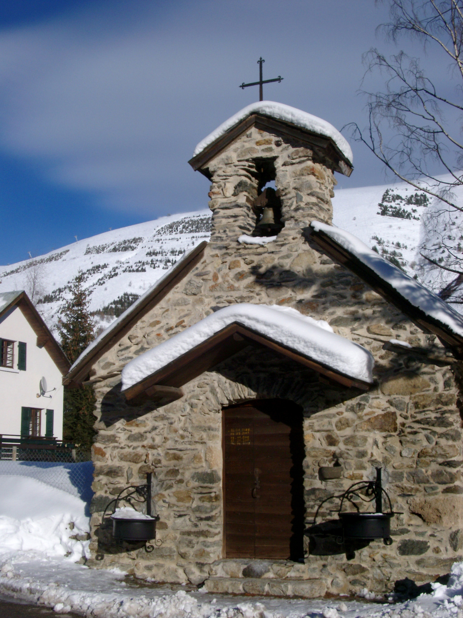 an image of Snows on a Small Historic Alpine Christian Church During Winter Season. Captured with Light Blue Gray Sky Background.