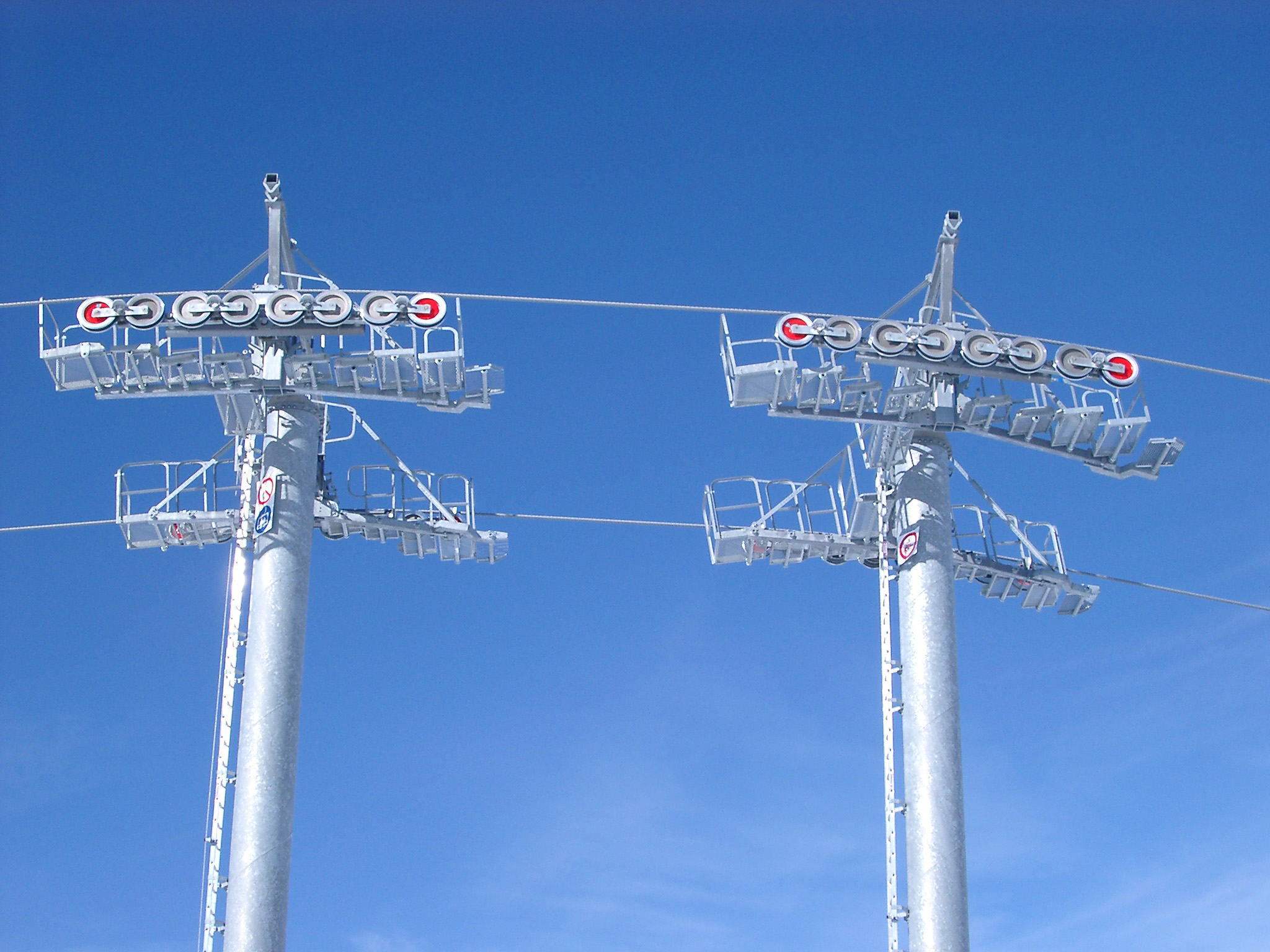 an image of Detail of an alpine chair lift showing the towers and cables with the pulleys against a clear blue sunny winter sky