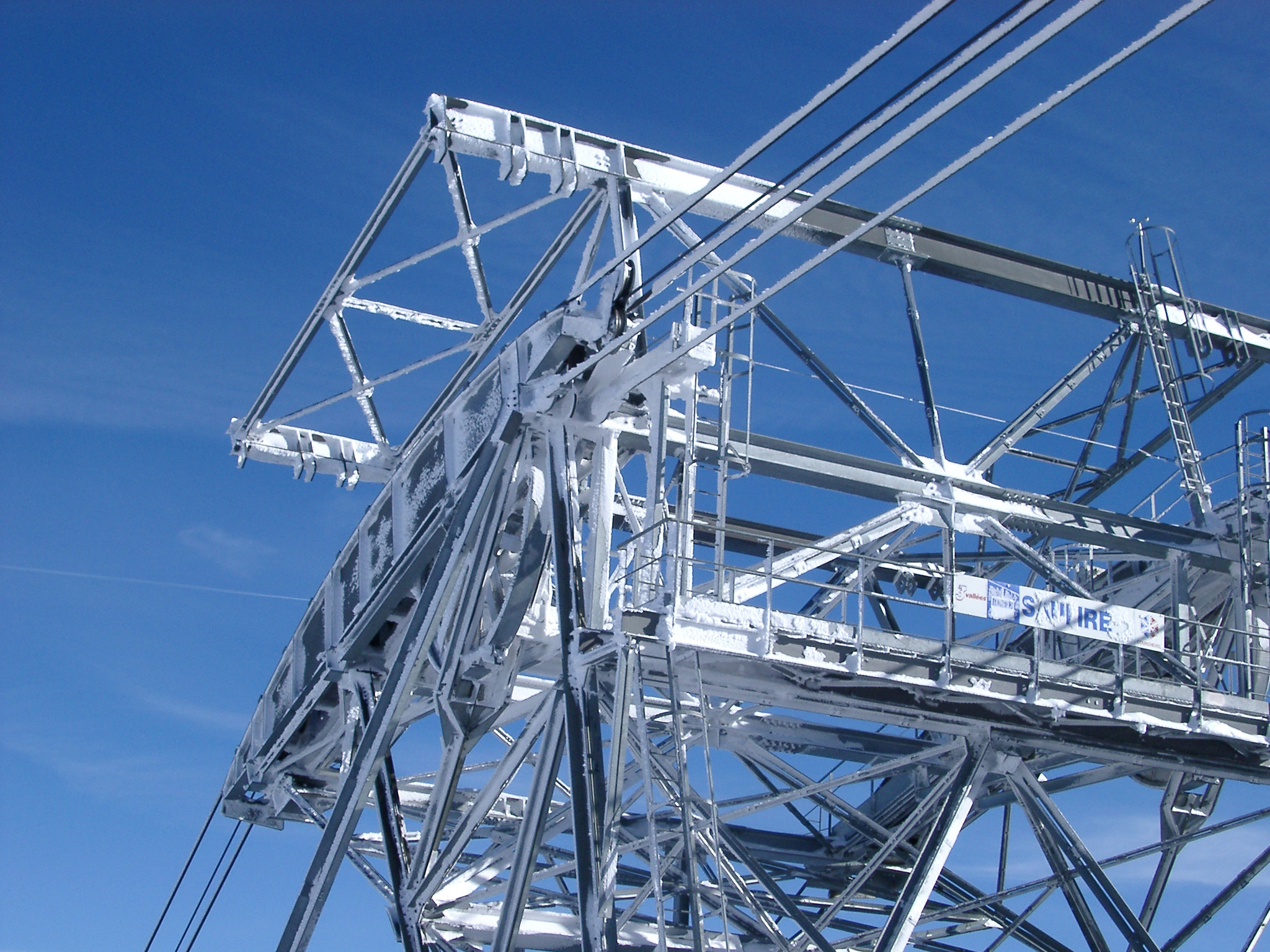 an image of Detail of a cable car tower showing the steel framework and cables on a ski slope against a sunny blue sky